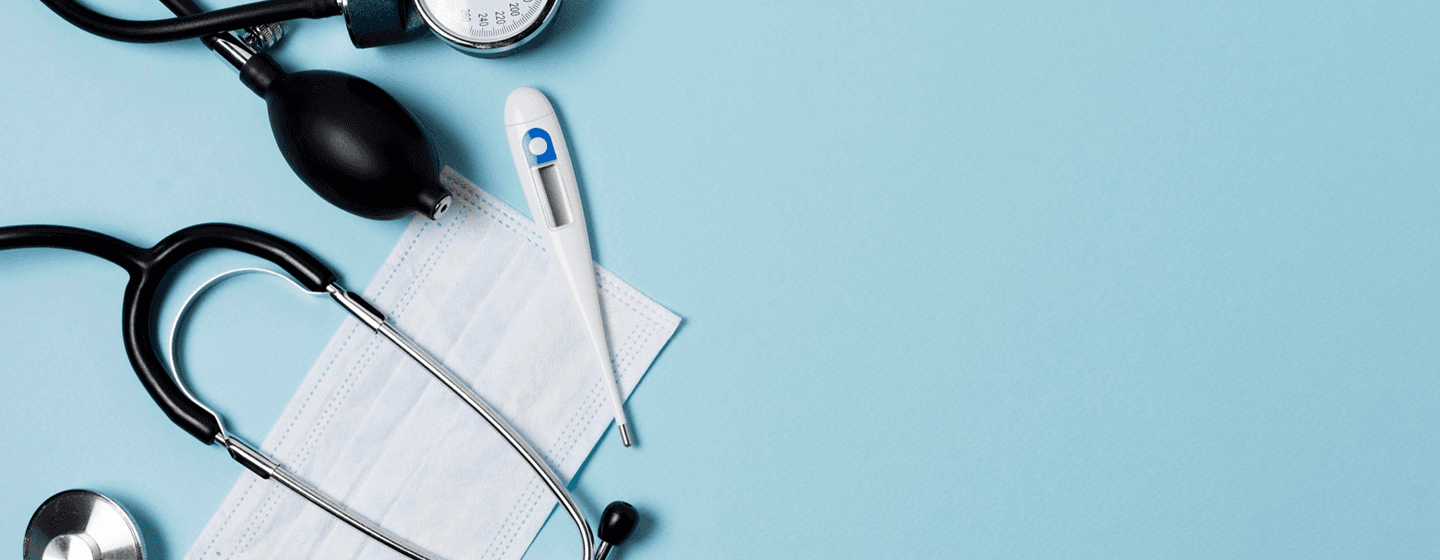A stethoscope, blood-pressure monitor, digital thermometer and surgical mask arranged on a blue background